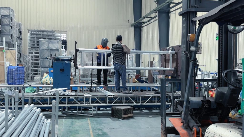 Worker welding aluminum spigot truss system components during stage truss manufacturing process